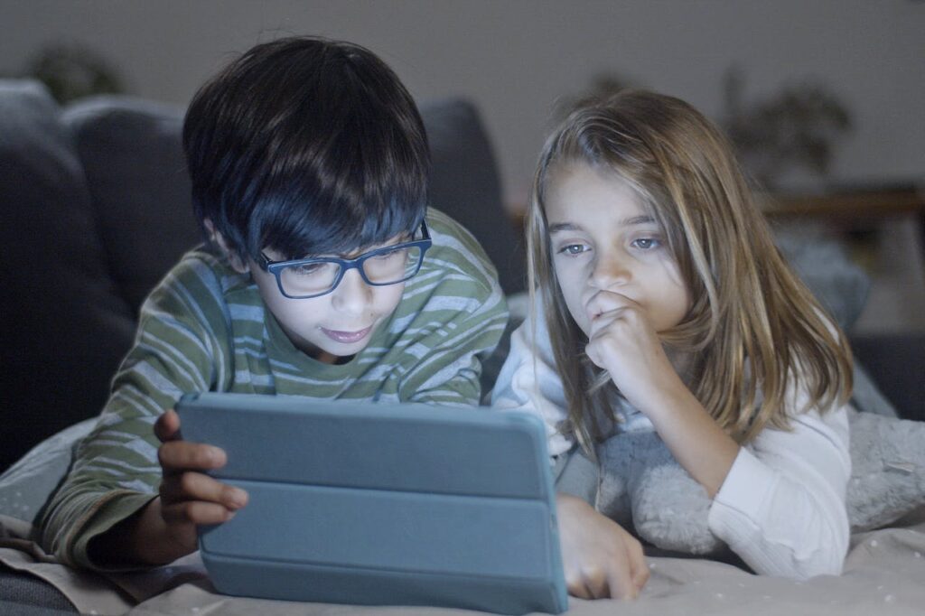 A boy and a girl are lying on the bed, watching something on their iPad.